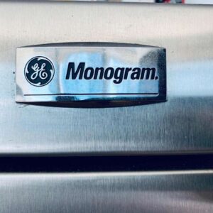 A close-up of a stainless steel refrigerator door with a circular monogram logo in the center.
